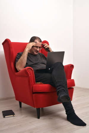 Middle-aged man with a laptop on his lap sed in a red armchair. The man is holding two smartphonesの写真素材