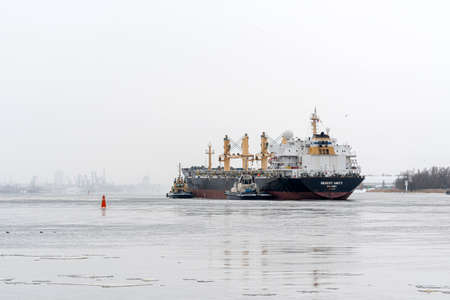 Riga, Latvia - February 9, 2021: multiple tugboats work in tandem to assist cargo ship to maneuver...