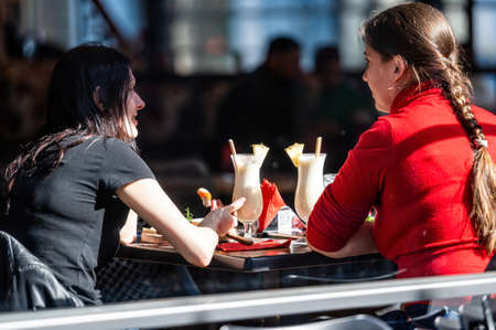 Riga, Latvia - April 2, 2022: Young women sitting in a cafe drinking coffee and talking on a sunny dayのeditorial素材