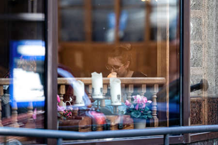 Riga, Latvia - April 2, 2022: A woman at a table in a restaurant. View through the window. Street reflections in the glass.のeditorial素材