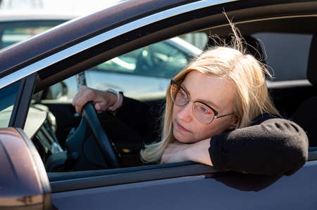thoughtful middle-aged blonde in glasses sits behind the wheel of a car, view through the window