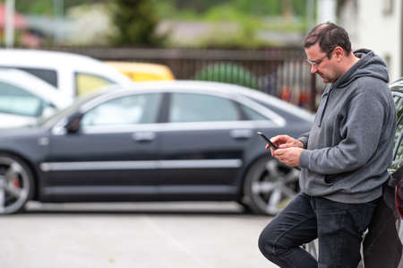 man with smartphone standing next to the car, using mobile app for online communication, shpping