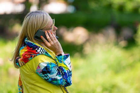 brightly dressed blonde senior woman talking on smartphone in garden, online communication conceptの写真素材
