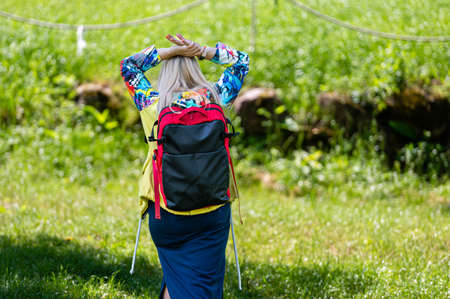 blonde senior woman in bright clothes with a backpack in the park on a sunny day, rear viewの写真素材