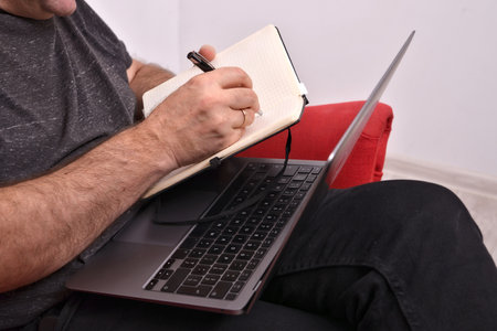 a man with a laptop on his lap taking notes in a notebook, closeup, online business analysis concept