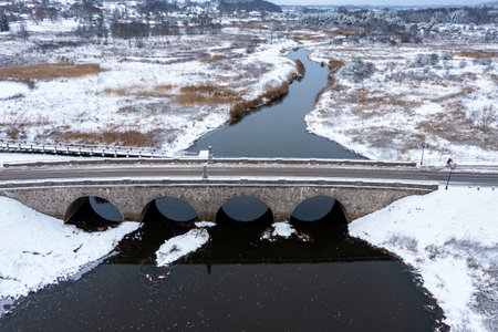 An old stone bridge with arches over the Abava river on a snowy winter day, Kandava,  aerial view