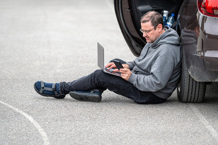 middle-aged man sitting on the asphalt by the car and working on a laptop, working remotely, travel