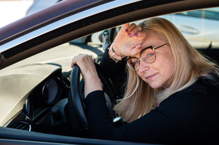 thoughtful middle-aged blonde in glasses sits behind the wheel of a car, view through the window