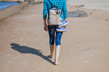 woman with backpack walking barefoot on beach during a sunny day, rear view, close-upの写真素材
