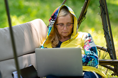 mature woman with laptop and documents working in garden on rocking couch, green home office conceptの写真素材