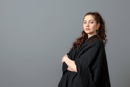 portrait of charming curly-haired teenager woman in a black cloak isolated on a gray backgroundの写真素材