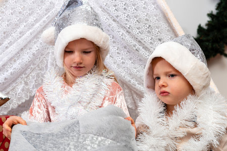 young children in silver Christmas hats sit between gift boxes and toys in a decorated house, Christmas time conceptの写真素材