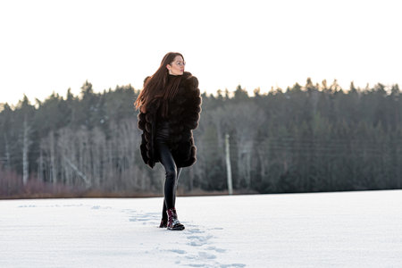 a cheerful woman in a warm fur coat walks across the open field leaving footprint in the snowの写真素材