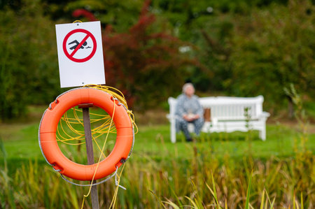 A sign indicates no swimming beside a lifebuoy near a peaceful garden bench.の写真素材