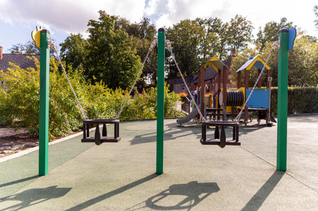 Swings stand empty in a bright and sunny playground with nearby play structures and trees.