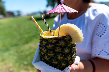 A person holds a vibrant pineapple drink adorned with colorful umbrellas in a sunny park.の写真素材