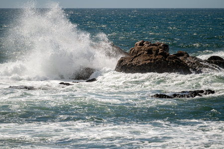 Powerful waves crash onto rocks along the coastline, creating a misty spray.の写真素材