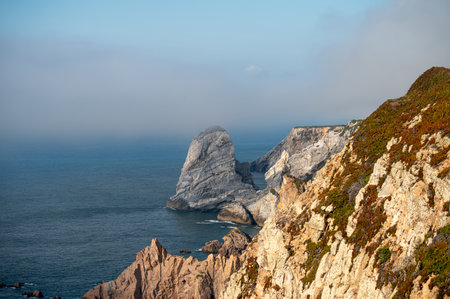 The sun casts a warm glow on rugged cliffs above tranquil waters with a misty backdrop.の写真素材
