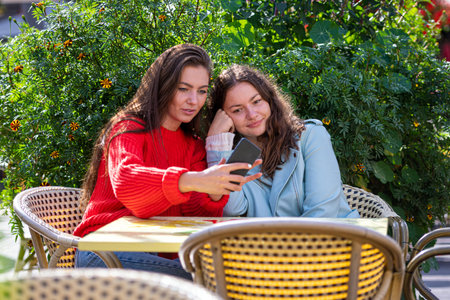 two young girls sit in a green cafe, chatting, laughing and taking pictures on a smartphoneの写真素材