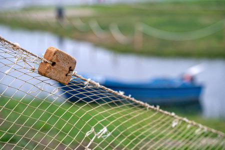 A wooden float hangs from a fishing net near a blue boat along a tranquil riverbank.の写真素材