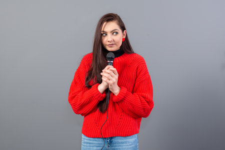 young, emotional brunette with a microphone dressed in a red sweater sings karaoke or says a speech, isolated on grayの写真素材