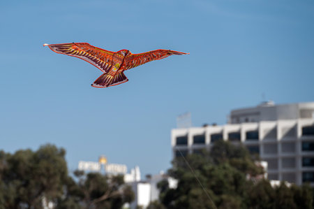 A realistic bird-shaped kite gliding against a clear blue sky with buildings in the background