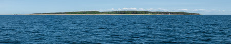Coastline features lush trees, tranquil blue sea, and clear skies under bright sunlight.の写真素材