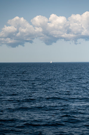 A small sailboat sails peacefully across expansive blue waters beneath a cloud-filled sky.の写真素材