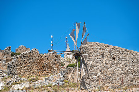 A traditional windmill overlooks ancient stone ruins under a bright blue sky in Crete, Greece.の写真素材