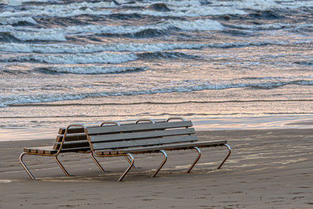 Benches sit empty on the beach as waves softly roll in during sunset.の写真素材