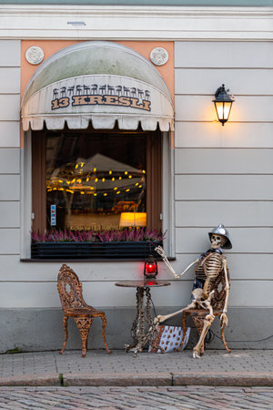Riga, Latvia - November 16, 2018: A skeleton dressed in a hat and scarf sits at a quaint cafe table outside.のeditorial素材