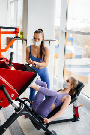 Two young beautiful girls exercise in the gym wearing masks during the pandemic. Social distancing in public places.の写真素材