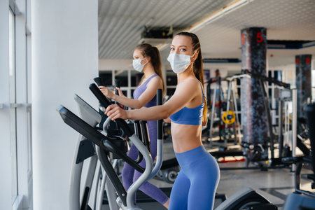 Two young beautiful girls exercise in the gym wearing masks during the pandemic. Social distancing in public places.の写真素材