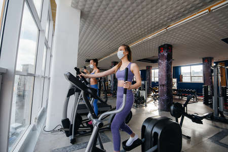 Two young beautiful girls exercise in the gym wearing masks during the pandemic. Social distancing...