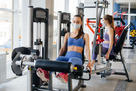 Two young beautiful girls exercise in the gym wearing masks during the pandemic. Social distancing...