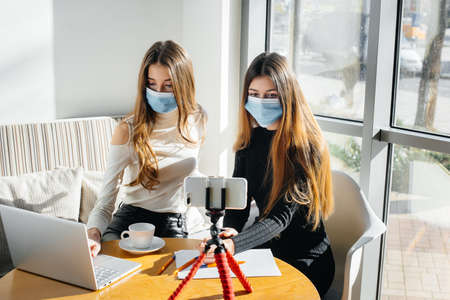 Two young girls sit in a cafe in masks and lead a video blog. Communication to the camera.