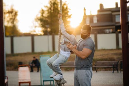 A father helps his son climb a rope on a sports field in masks during sunset. Healthy parenting and healthy lifestyleの写真素材