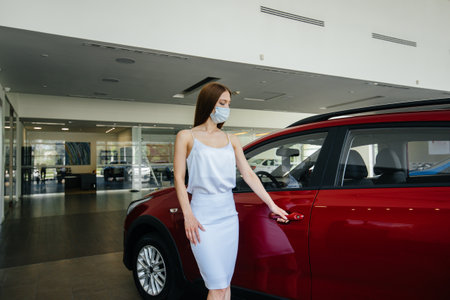 A young pretty girl inspects a new car at a car dealership in a mask during the pandemic. The sale...