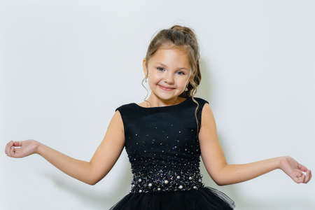 A little cute girl in a black dress sits and poses on a white background.の写真素材