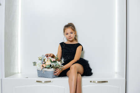 A little cute girl in a black dress is sitting and smiling with a bouquet of flowers.