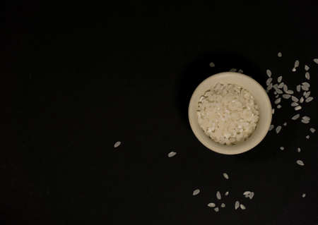 White uncooked rice in a bowl with scattered grains nearby on a black backgroundの写真素材