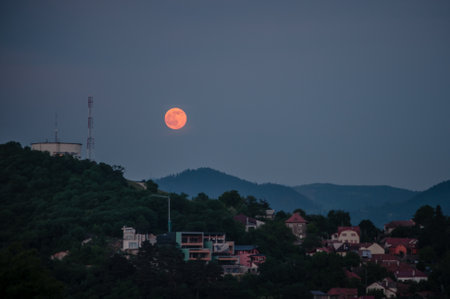 A stunning image of a super moon rising over a city nestled on a hill, with a serene mountain backdrop at twilight. The glowing moon and tranquil evening atmosphere create a picturesque and celestial scene, perfect for night photography and astronomy enthusiasts.の写真素材