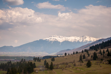 A serene spring landscape showcasing the Bucegi Mountains in the distance, featuring snow-capped peaks rising above lush green fields. The scene is complemented by dynamic cloud formations, creating a picturesque and tranquil setting ideal for nature lovers and hikers.の写真素材