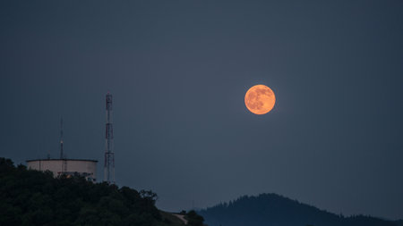 A striking image of a super moon rising above a hilltop water tank and communication tower, set against a tranquil twilight sky. The glowing moon adds a serene and atmospheric touch to this blend of nature and industrial elements, perfect for night photography and astronomy enthusiasts.の写真素材
