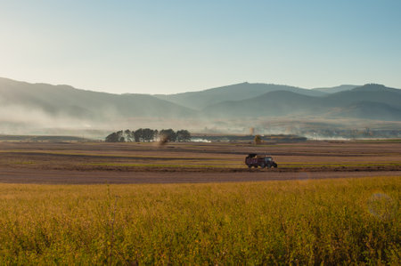 A picturesque rural scene capturing a tractor working on agricultural land during a misty morning....