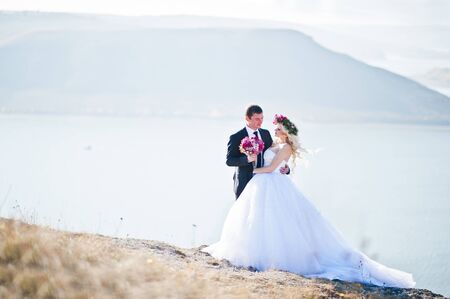 Charming bride in a wreath and elegant groom on landscapes of mountains, water and blue sky at sunny weatherの写真素材