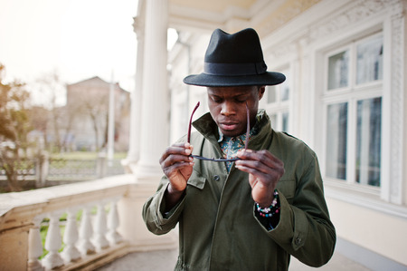 Close up fashion portrait of black african american man on green coat cloak and black hat with sunglasses stay background yellow mansionの写真素材