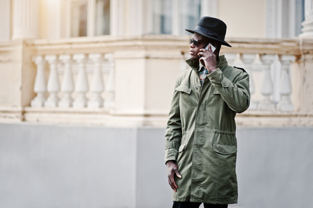 Fashion portrait of black african american man on green coat cloak and black hat,talking on the mobile phone background yellow mansionの写真素材