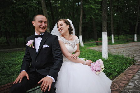 Cheerful wedding couple sitting on bench leaning backs to each otherの写真素材