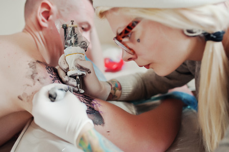 Young blonde woman master tattooist in glasses and hat with tattoo machine at hands make tattoo Dreamcatcher at hand of man. Close upの写真素材